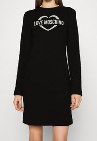 Svart klänning med långa ärmar och texten "LOVE MOSCHINO" inuti en hjärtform på bröstet, bärs av en stående person mot en enfärgad bakgrund.