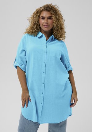 Vrouw met krullend blond haar, gekleed in een lichtblauwe jurk met knopen en opgerolde mouwen, gecombineerd met lichtblauwe jeans, staand tegen een effen achtergrond.