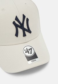 Casquette de baseball beige avec un logo brodé « NY » bleu marine. Caractéristiques d'une visière courbée et d'une étiquette de marque indiquant « ’47 OSFA ». Tissu texturé.