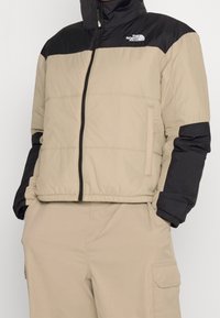 Beige pufferjacka med svarta axlar, hel dragkedja, två sidofickor och ribbade ärmslut. Tyget har en slät textur och en lätt glans.