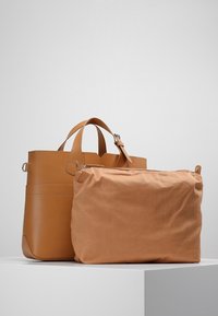 Tote bag van tan leer met een gestructureerd ontwerp en korte handvatten, naast een zachte, tan stoffen tas met een ritssluiting.