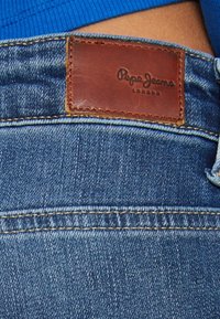 Close-up of blue denim jeans with brown leather "Pepe Jeans London" patch above pocket, worn with a blue ribbed top.