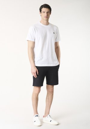 Uomo in piedi che indossa una maglietta bianca a maniche corte, pantaloncini neri e scarpe da ginnastica bianche su uno sfondo chiaro e uniforme.