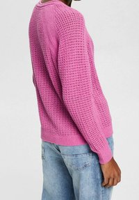 Pullover rosa lavorato a maglia con un design morbido e testurizzato, polsini e orlo a coste, indossato sopra jeans in denim azzurri chiari.