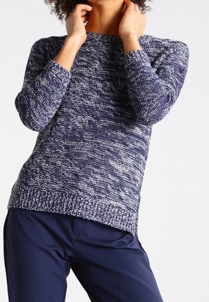 Personne portant un pull en maille à manches longues bleu et blanc chiné et un pantalon bleu marine, ajustant ses cheveux autour du cou.
