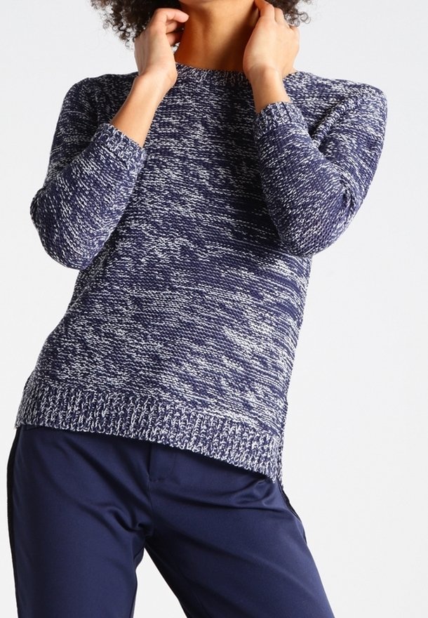 Personne portant un pull en maille à manches longues bleu et blanc chiné et un pantalon bleu marine, ajustant ses cheveux autour du cou.