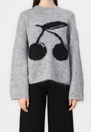 Žena oblečená v šedém chlupatém svetru s velkým černým motivem třešně na přední straně, doplněném černými kalhotami.