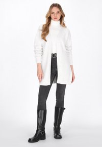 Cardigan bianco lavorato a maglia con maniche lunghe, apertura frontale, orlo a coste, abbinato a jeans skinny neri e stivali alti neri in pelle.