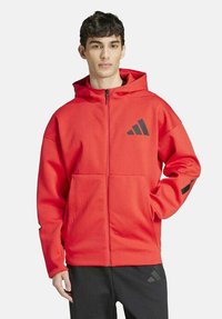 Rode zip-up hoodie van textiel met een capuchon. Heeft een zwart logo en accenten op de mouwen. Voorkwam pockets zichtbaar.