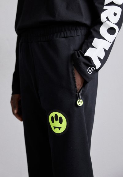 Black jogger pants with elastic waistband, featuring a large green logo design and a zippered pocket accented with a green pull tab.