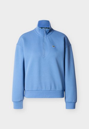 Hellblauer Pullover mit hohem Kragen, halbem Reißverschluss vorne und gerippten Bündchen. Verfügt über ein kleines, gesticktes Logo auf der Brust. Hergestellt aus weichem Stoff.