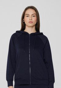 Sudadera de cremallera en color marino, hecha de tela suave, que presenta una capucha con cordón, mangas raglán, y puños y dobladillo de canalé. Textura suave, diseño minimalista.