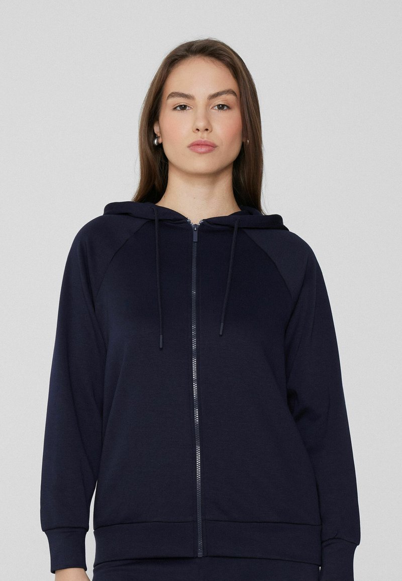 Sudadera de cremallera en color marino, hecha de tela suave, que presenta una capucha con cordón, mangas raglán, y puños y dobladillo de canalé. Textura suave, diseño minimalista.