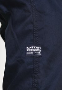 Námořnická modrá denimová látka s vertikální texturou; má na straně vyšitou nášivku G-Star Originals v černé a bílé barvě.