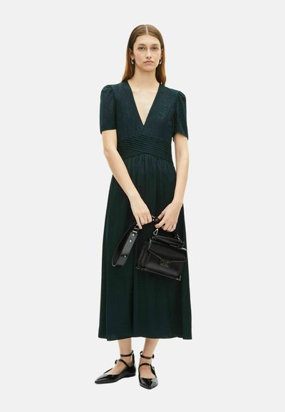 Frau, die ein dunkelgrünes Midikleid mit kurzen Ärmeln trägt, eine schwarze Lederhandtasche hält und schwarze Ballerinas mit Knöchelriemen trägt.