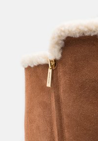 Brown suede ankle boot with cream shearling lining, side zipper featuring a gold accent tag, textured surface, and stitched detailing.