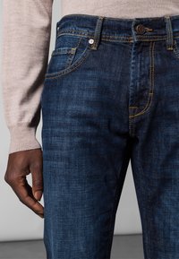 Dark blue jeans made of denim with yellow stitching, five-pocket design, and a button fly, paired with a light beige sweater.