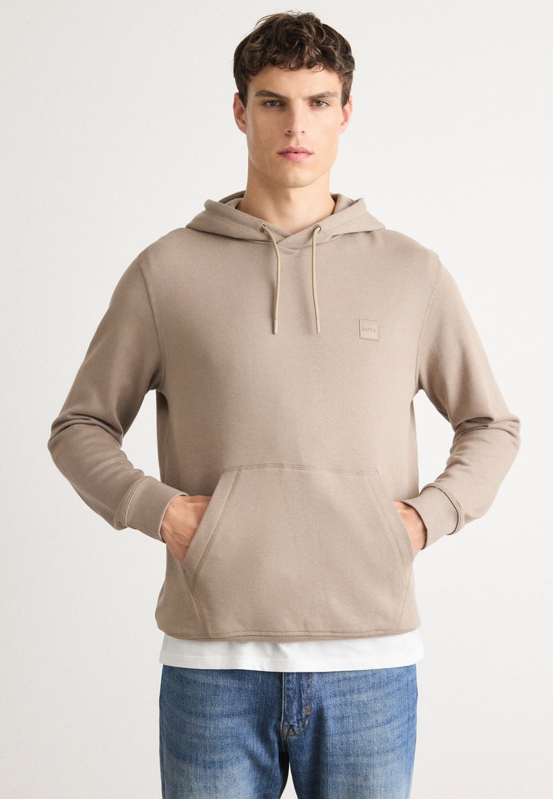 Jonge man in een beige hoodie met een kangoeroezak en blauwe jeans, staand tegen een effen witte achtergrond.