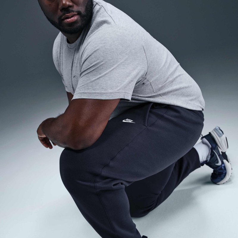 Uomo inginocchiato su un ginocchio, indossa una t-shirt grigia, pantaloni della tuta Nike neri, calzini bianchi e scarpe sportive nere, guarda di lato.