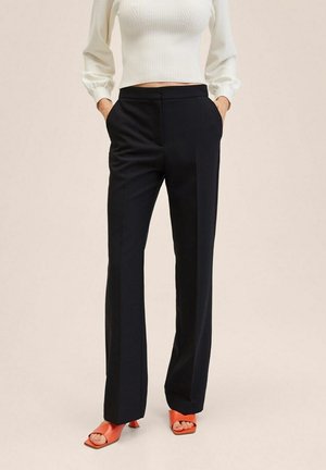 Frau trägt schwarze gerade geschnittene Hosen, weißes, geripptes, langärmeliges Croptop und orangefarbene offene High Heels, Hände in den Taschen.