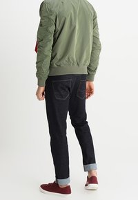 Veste bomber verte olive avec des poignets côtelés, associée à un jean bleu foncé et des baskets bordeaux. La veste comporte une accent rouge sur la manche.