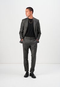Costum gri cu model, cu croială slim, dintr-un material texturat, asortat cu o cămașă neagră și pantofi negri, având un lanț auriu subtil.