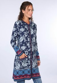 Marineblauer Mantel mit Blumenmuster und Kapuze, ausgestattet mit roten und blauen Akzenten, Knopfverschluss und Fronttaschen. Hergestellt aus weichem, strukturiertem Stoff.