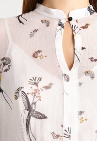 White, lightweight blouse with a keyhole neckline, featuring floral and butterfly patterns in black, orange, and yellow accents. Textured fabric.