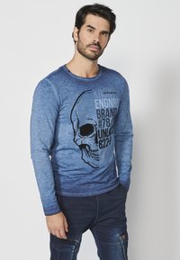 Majica s dugim rukavima u plavom gradijentu s crnom slikom lubanje i tekstom. Izrađena od mekanog materijala, s okruglim izrezom i slim fit dizajnom.
