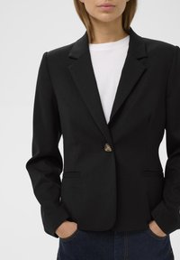 Fekete blézer, igényes szabású, egysoros gombolású, és kieső reverrel. Simító anyagból készült, két elülső zsebbel. Fehér póló alatta.