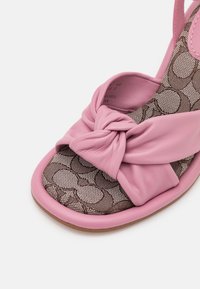 Sandale rose à bout ouvert avec bride en cuir nouée et semelle intérieure marron à motifs sur fond blanc.