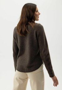 Brauner Wollpullover mit weicher Textur, langen Ärmeln und seitlichen Schlitzen. Kombiniert mit beigen Hosen, von hinten in einem neutralen Hintergrund gezeigt.