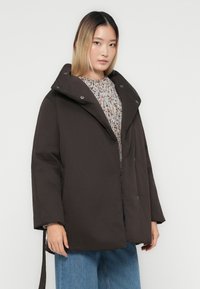 Ung kvinne iført en mørk overdimensjonert dunjakke over en flerfarget strikkegenser og blå jeans, stående foran en hvit bakgrunn.