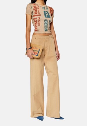 Kobieta w szerokich, beżowych spodniach i krótkim, graficznym t-shircie, trzyma beżową torebkę dopasowaną do spodni i nosi niebieskie buty na szpic.