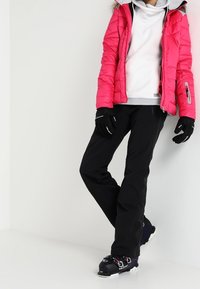Pink puffer jacket with a hood, gray lining; white long-sleeve shirt and black ski pants; black gloves; ski boots with colored accents.