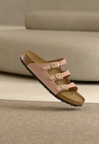 Ροζ σανδάλι με τρεις ράβδους, χρυσές αγκράφες, πάτο φελλού και μαύρη σόλα, τοποθετημένο δίπλα σε μπεζ καναπέ και χαλί.