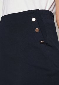 Navy blauwe rok van zacht materiaal, met een hoge taille en decoratieve rosegouden knopen aan de zijkant en een gladde textuur.