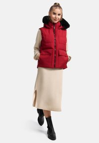 Gilet imbottito rosso con colletto e zip frontale, dotato di due grandi tasche anteriori. Indossato su un vestito beige a maniche lunghe e stivali neri.