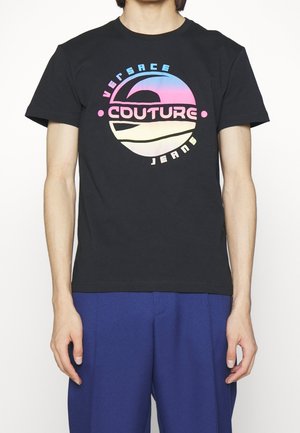 Czarny T-shirt z kolorowym, okrągłym logo "Versace Couture Jeans" noszony z niebieskimi spodniami, widok z przodu od szyi do bioder.