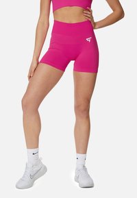 Dámske ružové športové šortky so zvýšeným pásom, hladkou textúrou a malým bielym logom na boku. Skombinované s bielymi teniskami a ponožkami.