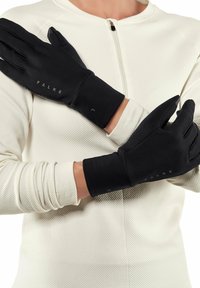 Person wearing black Falke gloves crossing arms over chest, dressed in a white textured long-sleeve top with a front zipper.
