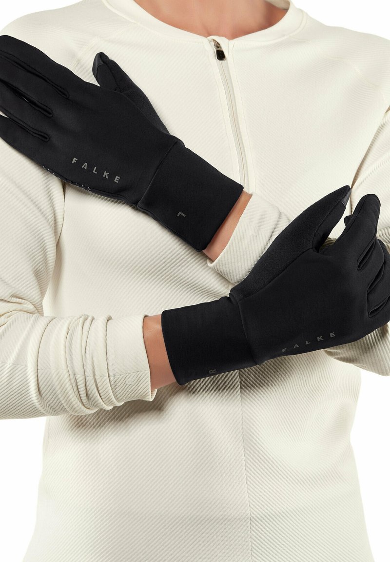 Person wearing black Falke gloves crossing arms over chest, dressed in a white textured long-sleeve top with a front zipper.
