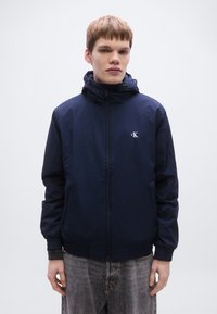 Μπουφάν ανεμοπαντελόνι σε ναυτικό μπλε χρώμα με κλείσιμο με φερμουάρ, κουκούλα, ελαστικές μανσέτες και μικρό λογότυπο στο στήθος. Το υλικό φαίνεται ελαφρύ και λείο.