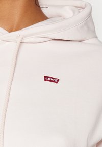 Haut à capuche rose clair en coton doux. Comprend un cordon de serrage pour la capuche et un petit patch logo rouge Levi's à l'avant.