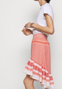 Röd mönstrad kjol med utställd nederkant, med en texturerad midja och volangkant i botten, lager av vita ränder och geometriska mönster.