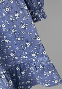 Blumenkleid aus hellblauem Stoff, mit weißen Blumen und grünen Blättern in einem wiederholten Muster. Elastische Details an der Taille und gewellter Saum.