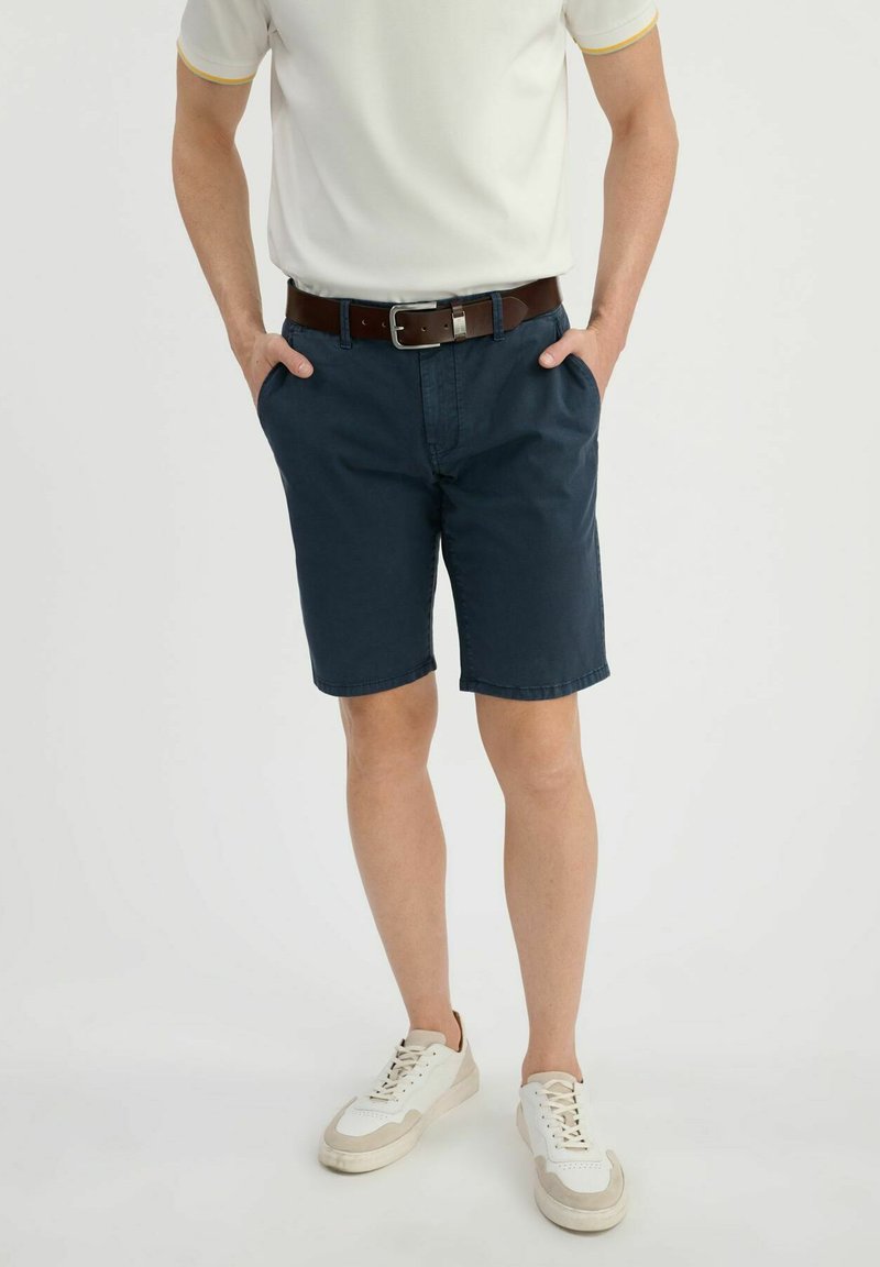 Granatowe szorty wykonane z bawełny, z płaskim przodem i dwoma kieszeniami po bokach. W zestawieniu z białym polo i brązowym paskiem. Białe sneakersy z beżowymi akcentami.