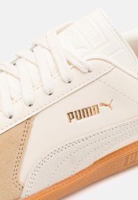 Weißer Ledersneaker mit hellbraunen Wildlederakzenten, goldfarbenem "PUMA"-Logo, flachen weißen Schnürsenkeln und einer strukturierten Gummisohle.