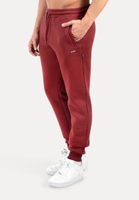 Bordeauxrote Jogginghose mit einem Taillenzugband, seitlichen Taschen und gerippten Bündchen, kombiniert mit weißen Sneakers. Glatte Stoffstruktur.