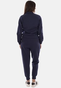Mujer de espaldas, vestida con un chándal azul marino y zapatillas blancas con detalles negros, sobre un fondo liso.